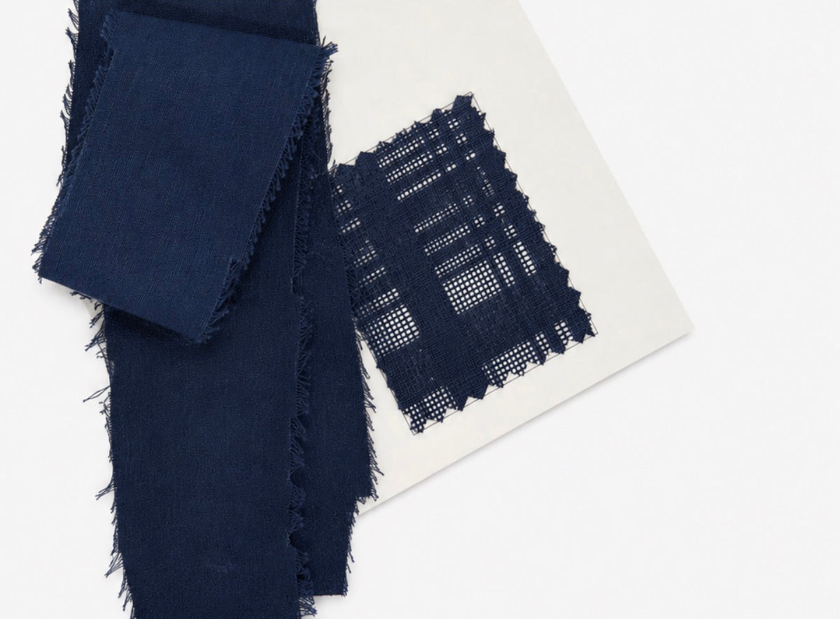 Blue woven Karami-ori fabric swatches with frayed edges, including a solid black square and a blue-and-white plaid-patterned square, displayed on a white card against a white background.