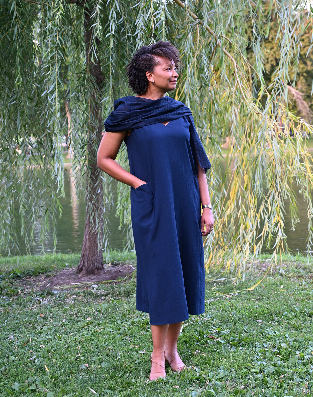 A woman wearing a navy blue PKTS Kelley midi dress with a matching Karami-ori cloth wrap is standing outdoors on a grassy lawn in front of a weeping willow tree by a body of water.