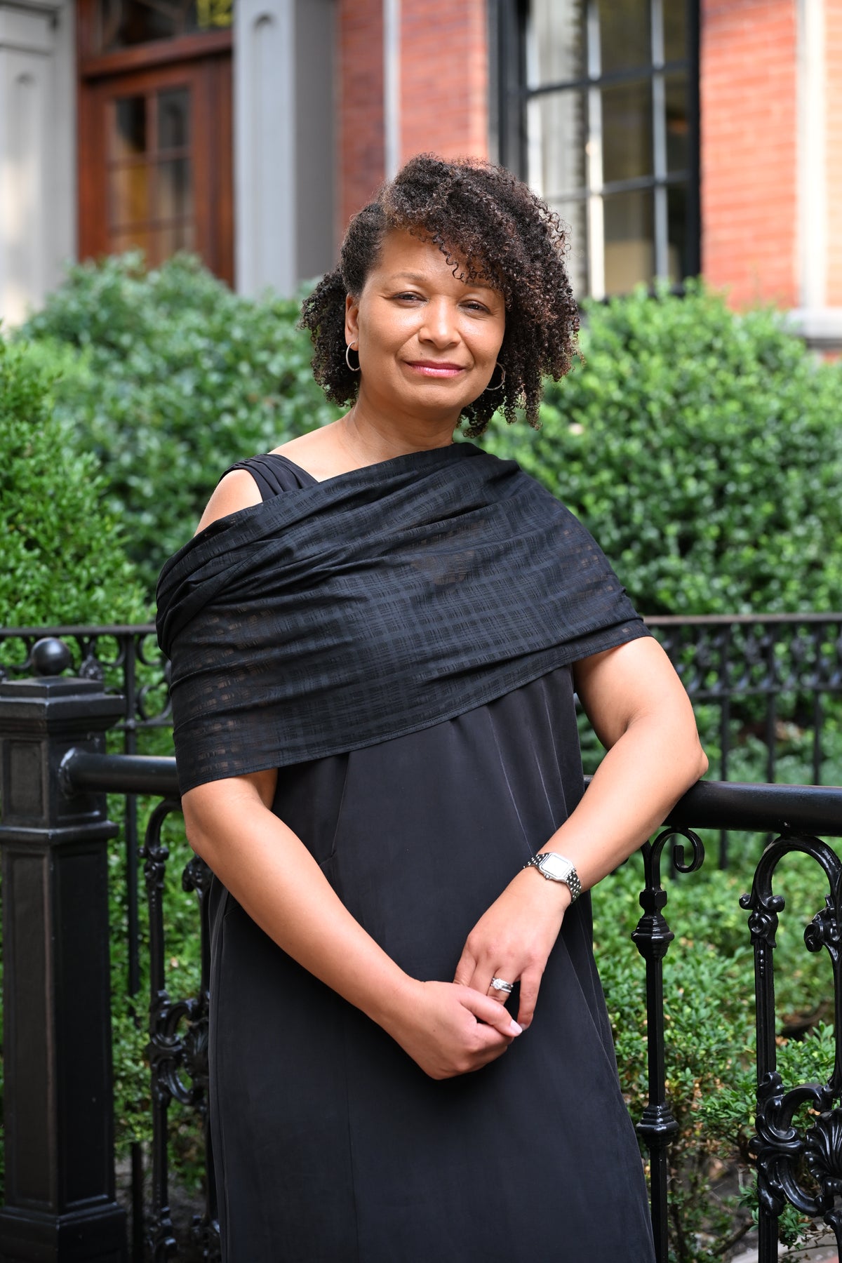 A woman with curly dark hair stands outdoors in front of a black wrought-iron fence and green hedges, wearing a black PKTS Kelly dress with an off-the-shoulder draped wrap.