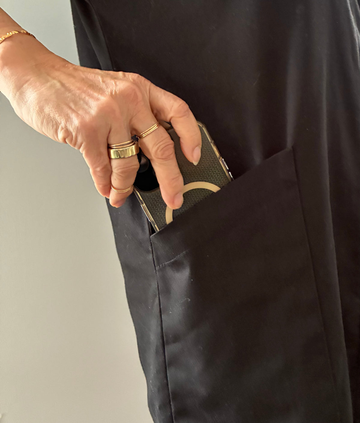 A close-up of a person's hand with gold rings reaching into a black fabric pocket, with a mobile phone partially visible inside.