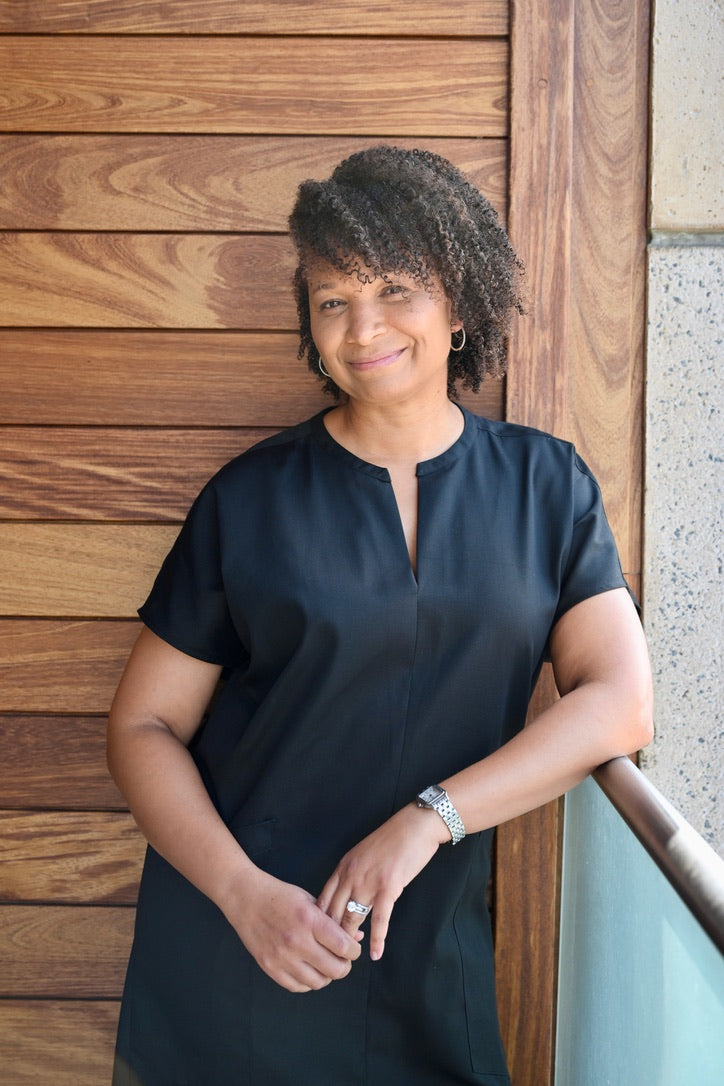 A woman with curly dark hair wearing a black short-sleeve dress with deep, functional pockets.