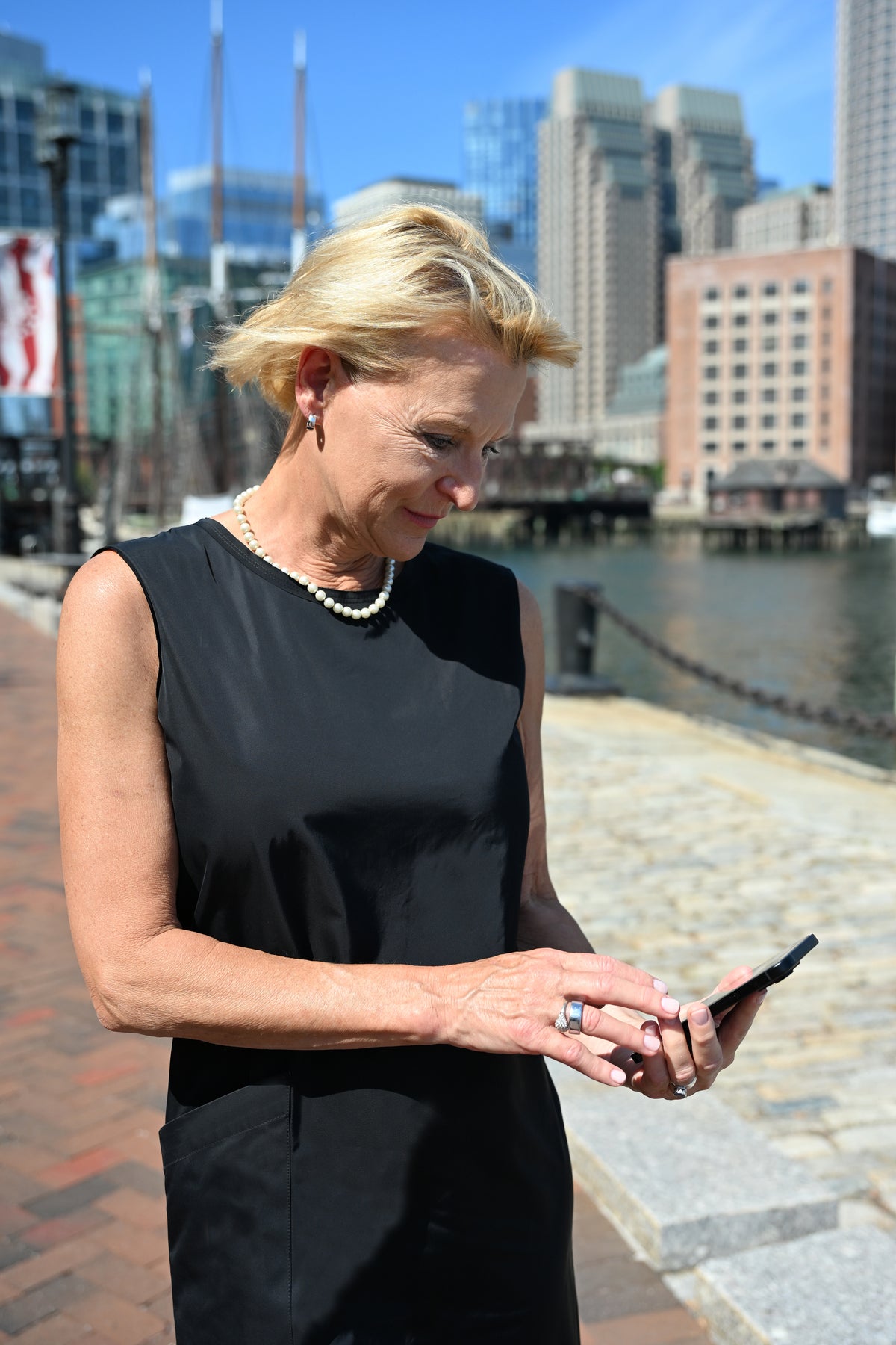 A blonde woman in a sleeveless black dress stands outdoors by a waterfront, looking down at a smartphone in her hands. Modern city buildings and a canal are visible in the background.