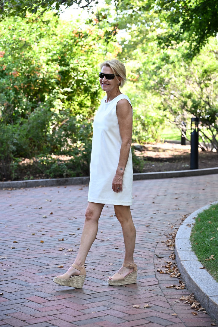 A woman wearing a sleeveless white shift dress and beige platform wedge sandals walks along a brick pathway in a park with green trees in the background.