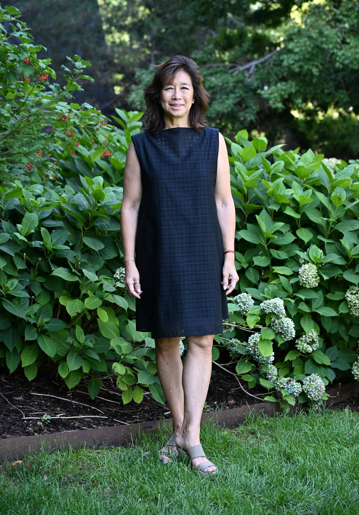 A woman in a black sleeveless PKTS Dana dress with a Karami-ori cloth overlay featuring a subtle grid pattern stands in a lush garden.