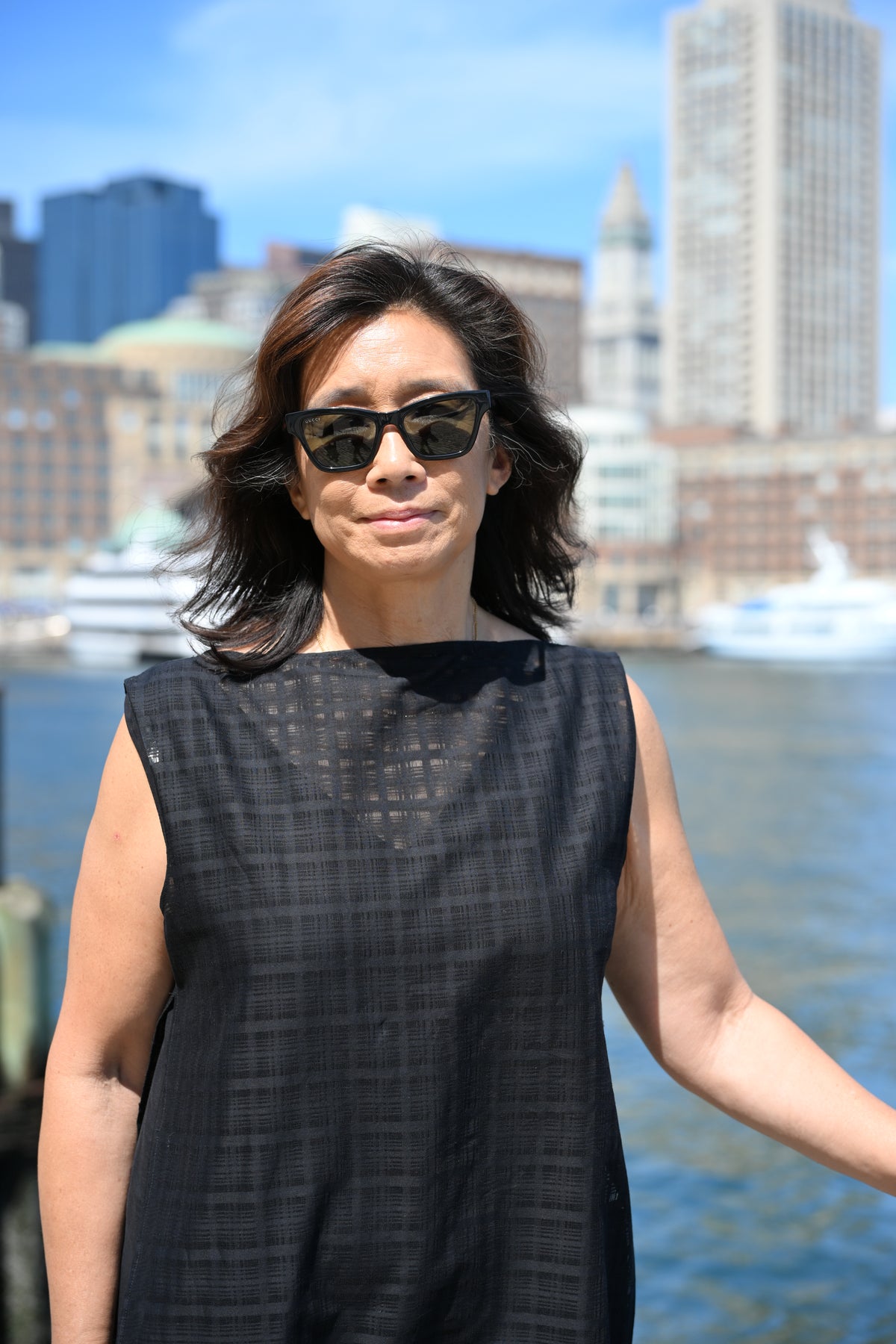 A woman wearing a sleeveless black PKTS Dana dress with a textured Karami-ori cloth overlay stands outdoors with a waterfront city skyline in the background.