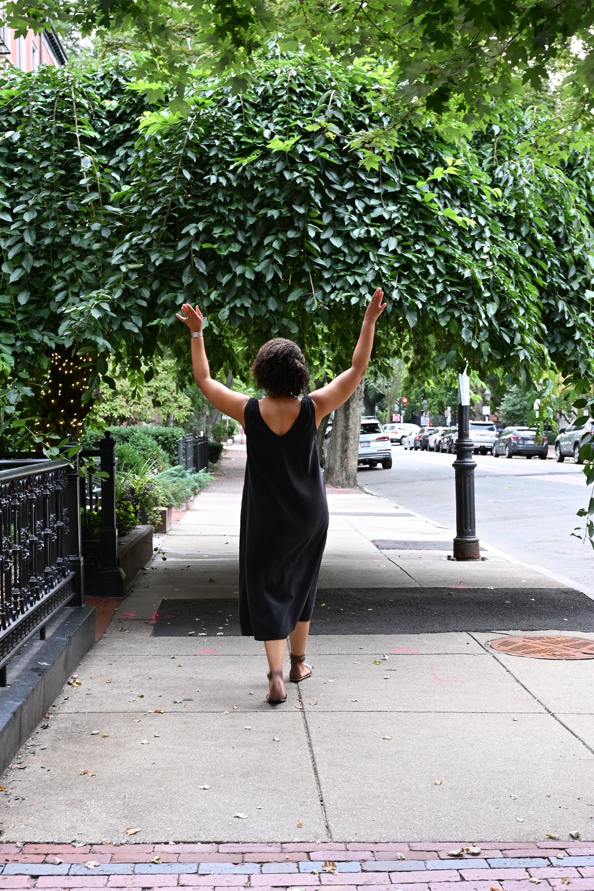 A woman wearing a black sleeveless PKTS Kelley midi dress walks away on a city sidewalk with her arms raised, surrounded by lush green trees and a street in the background.