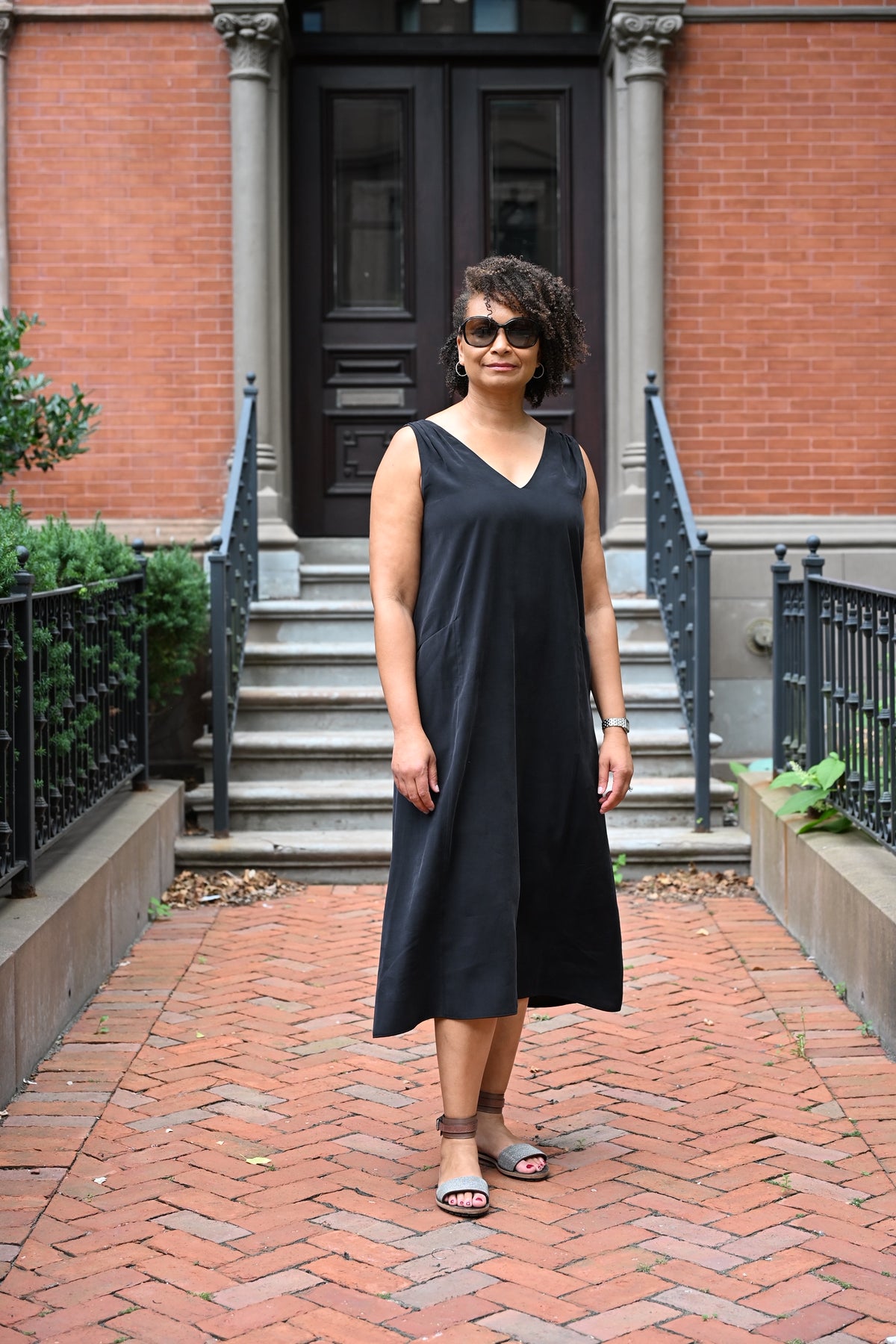 A woman wearing a sleeveless black PKTS Kelley midi dress and sunglasses stands on a brick walkway in front of a red brick building with stone steps.