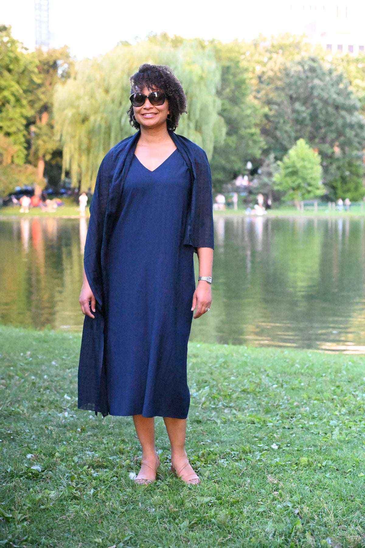 A woman wears a navy blue PKTS Kelley midi dress and Karami-ori cloth wrap stands on a grassy lawn in front of a park lake with trees in the background.
