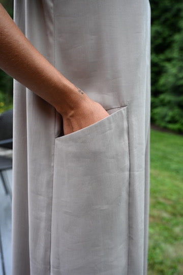 A close-up of a person wearing a sand PKTS Kelley dress with a deep, functional side pocket, with their hand in the pocket, set against a blurred green outdoor background.