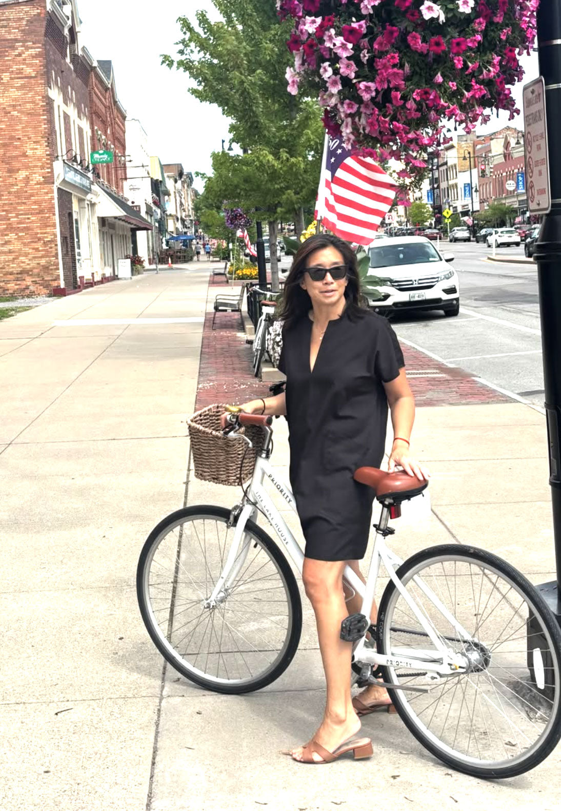 A woman wearing a black short-sleeve mini dress and sunglasses stands on a city sidewalk, straddling a white bicycle with a basket. The background shows a street scene with parked cars, trees, and hanging flower baskets.