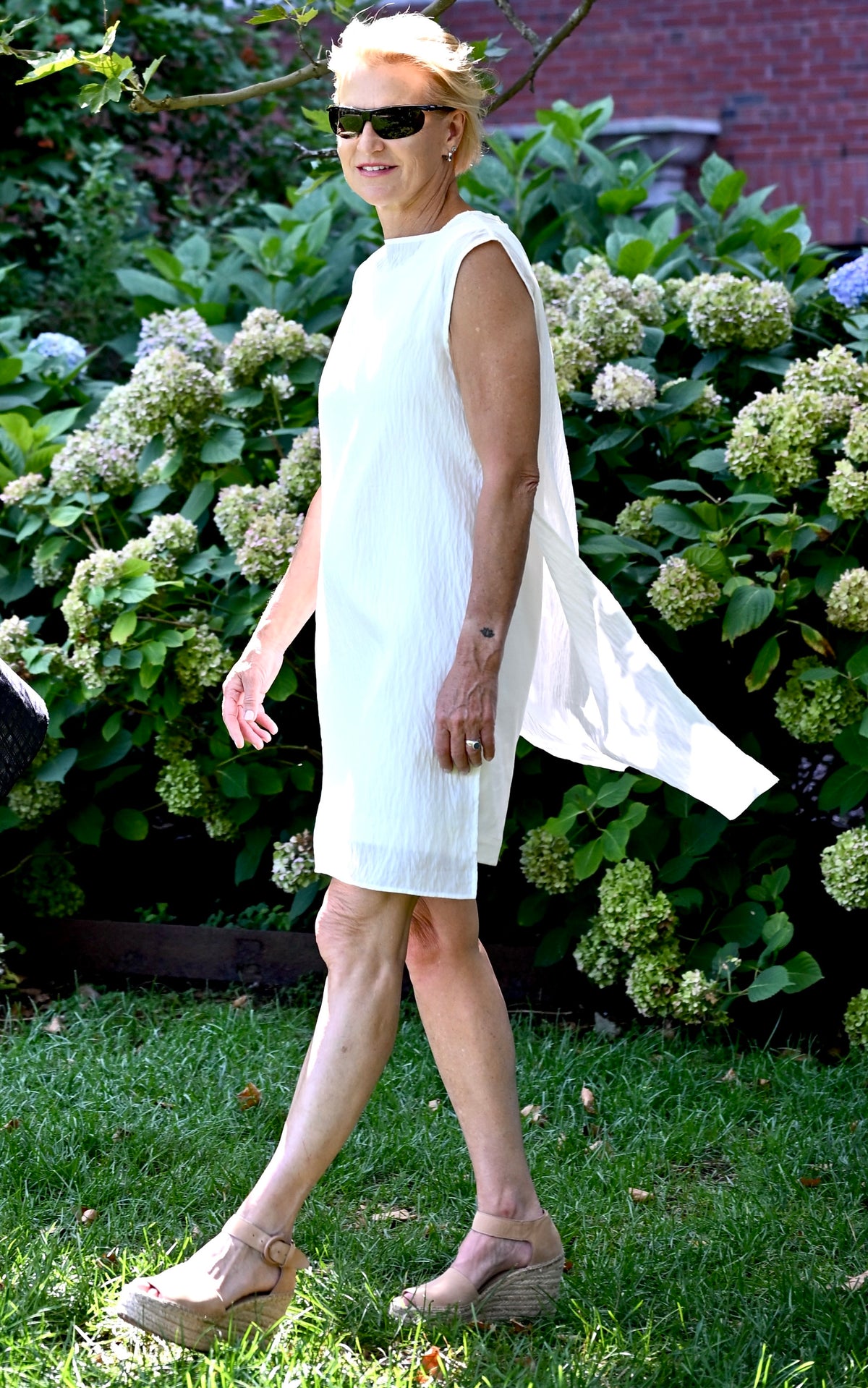 A woman walks outdoors in a sleeveless white PKTS Dana dress with a PKTS White Cloud overlay that highlights the overlay's flowing side drape, set against a backdrop of green grass and hydrangea bushes.