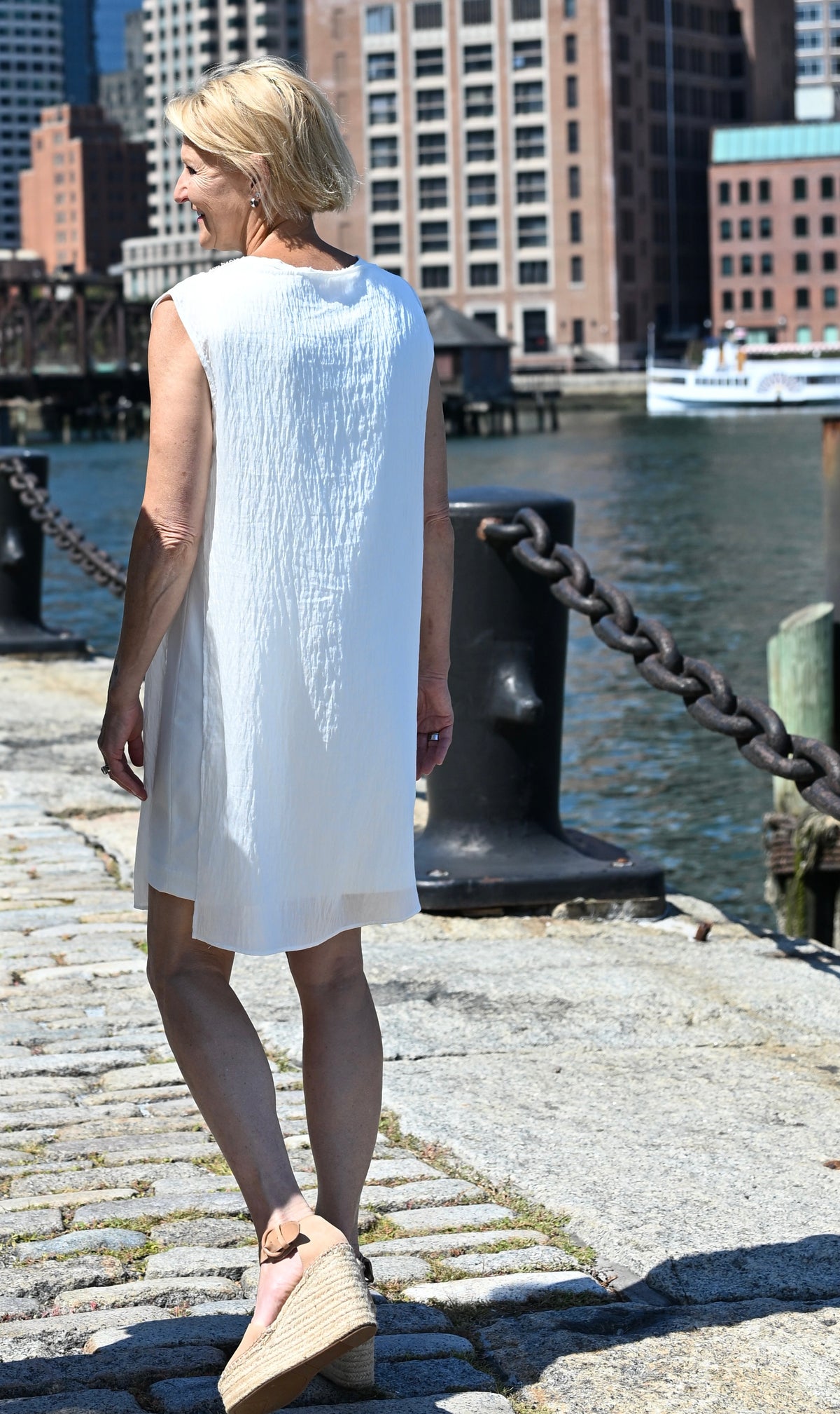 A woman wearing a sleeveless white PKTS Dana dress with a PKTS White Cloud overlay walks along a cobblestone waterfront path with a city harbor and buildings in the background.
