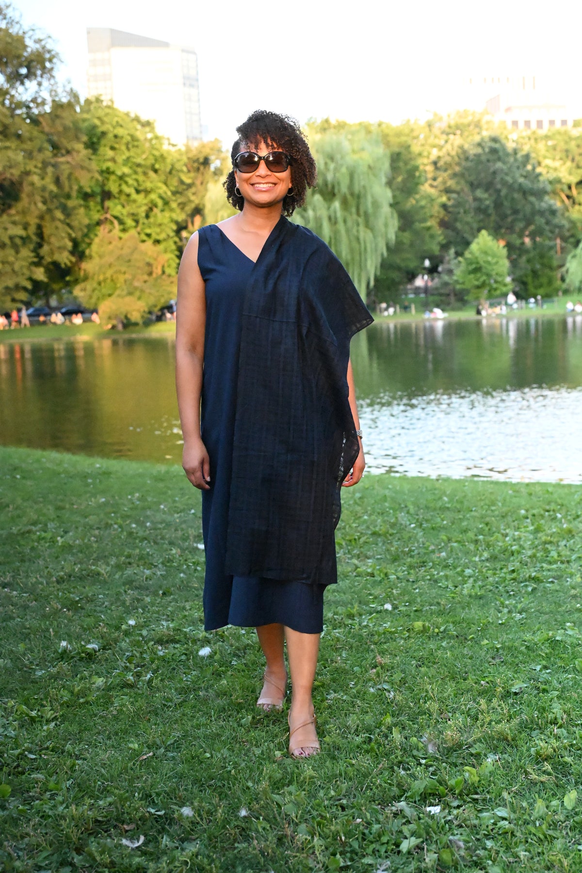 A woman wearing a dark navy blue sleeveless PKTS Kelley dress, draped with a one-sleeve Karami-ori cloth wrap, stands on a grassy lawn in front of a lake and trees.