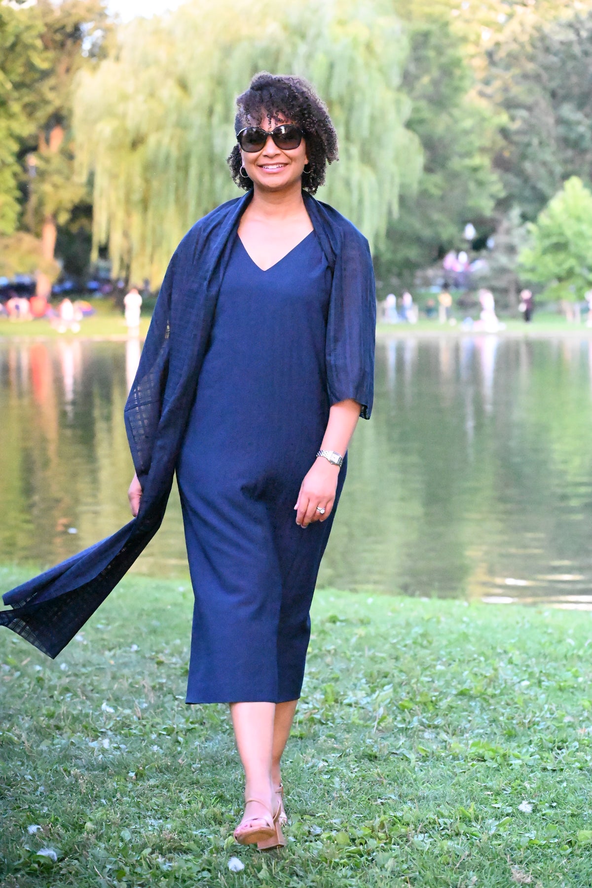 A woman wearing a navy blue PKTS Kelley midi dress and a draped Karami-ori cloth wrap walks along a grassy lakeside path with trees in the background.