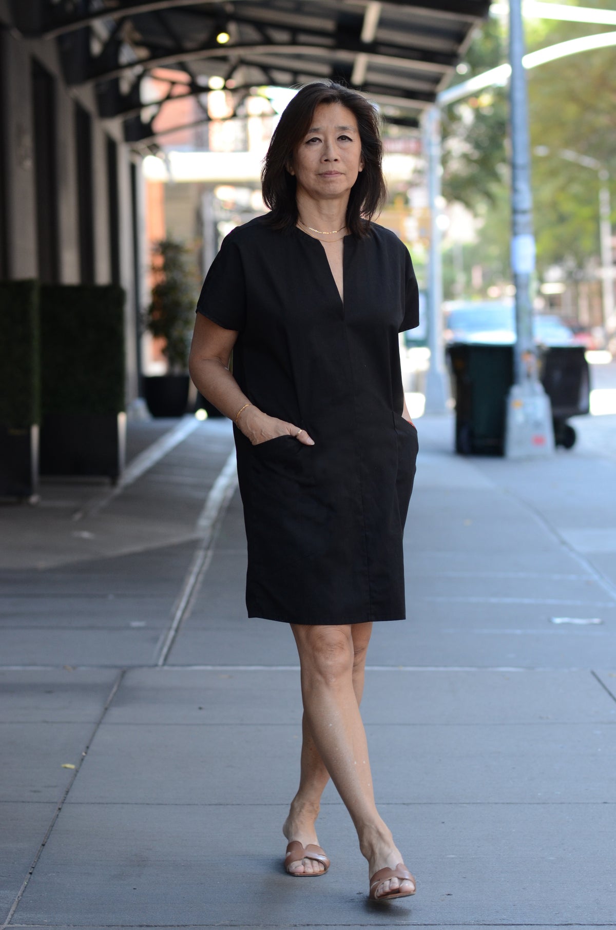 A woman wearing a black short-sleeve V-neck dress with deep, functional side pockets, standing on a city sidewalk under a covered walkway.