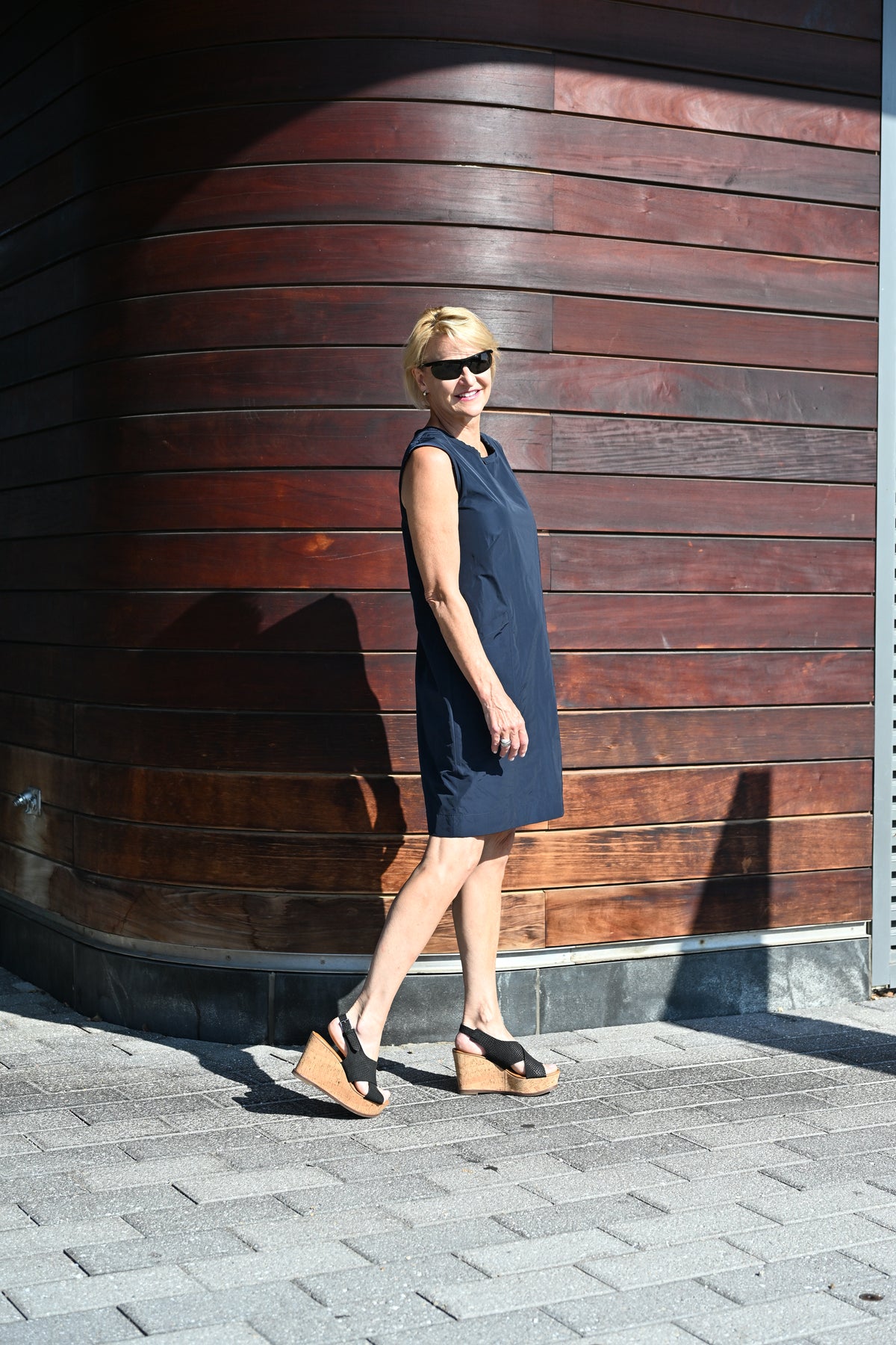 A woman wearing a sleeveless navy blue shift dress and black wedge sandals walks along a paved path in front of a dark brown wooden wall.
