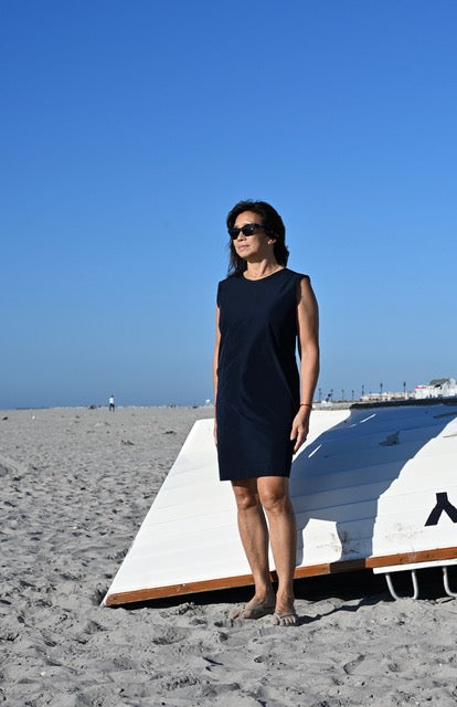 A woman wearing a sleeveless dark navy blue dress and sunglasses stands on a sandy beach next to a white boat hull under a clear blue sky.