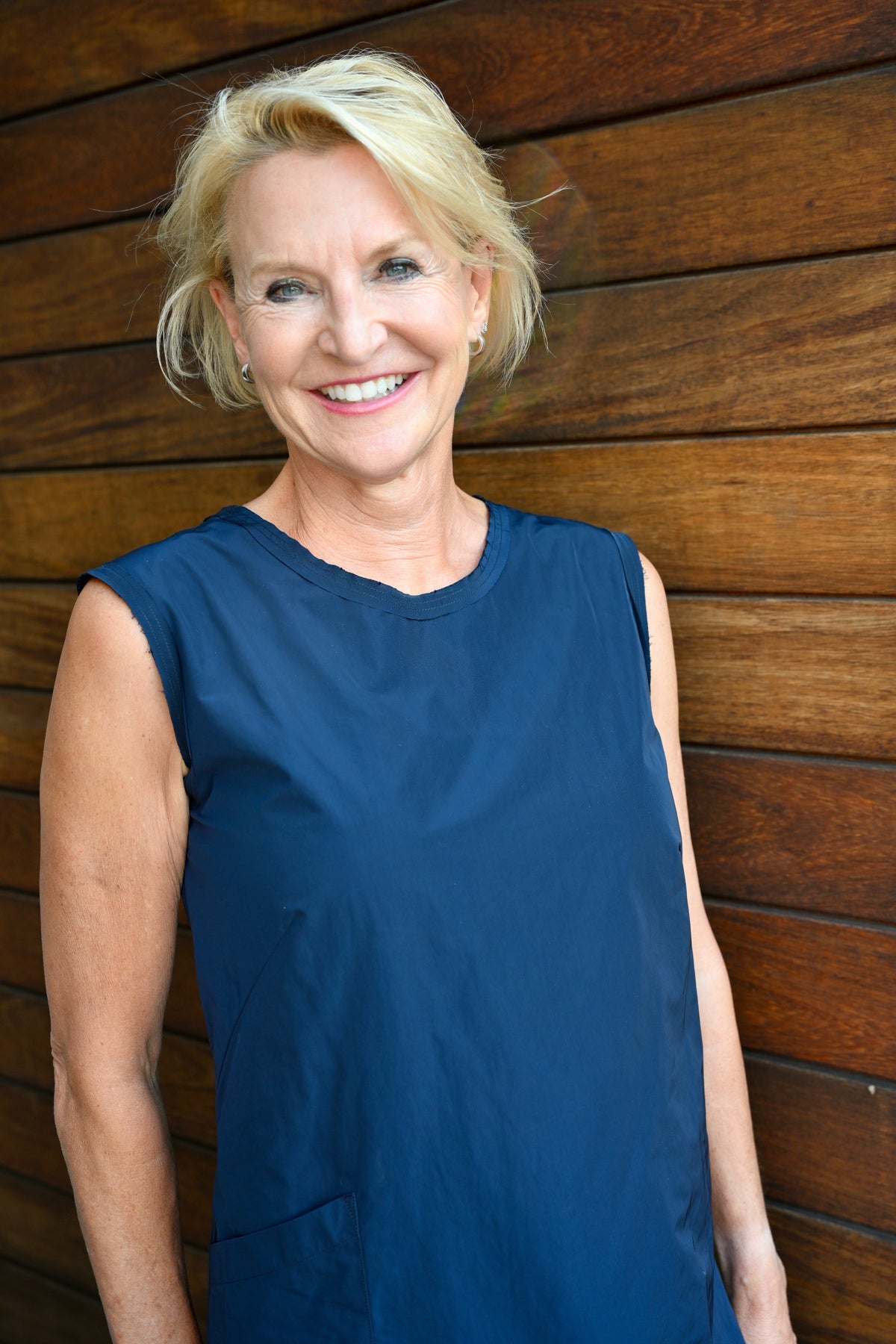 A smiling blonde woman wearing a sleeveless navy blue top, standing against a brown wooden plank wall.