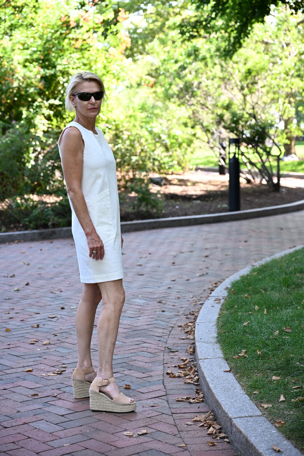 A woman wearing a sleeveless white shift dress and beige platform wedge sandals stands on a brick pathway in a park with green trees in the background with a serious expression.