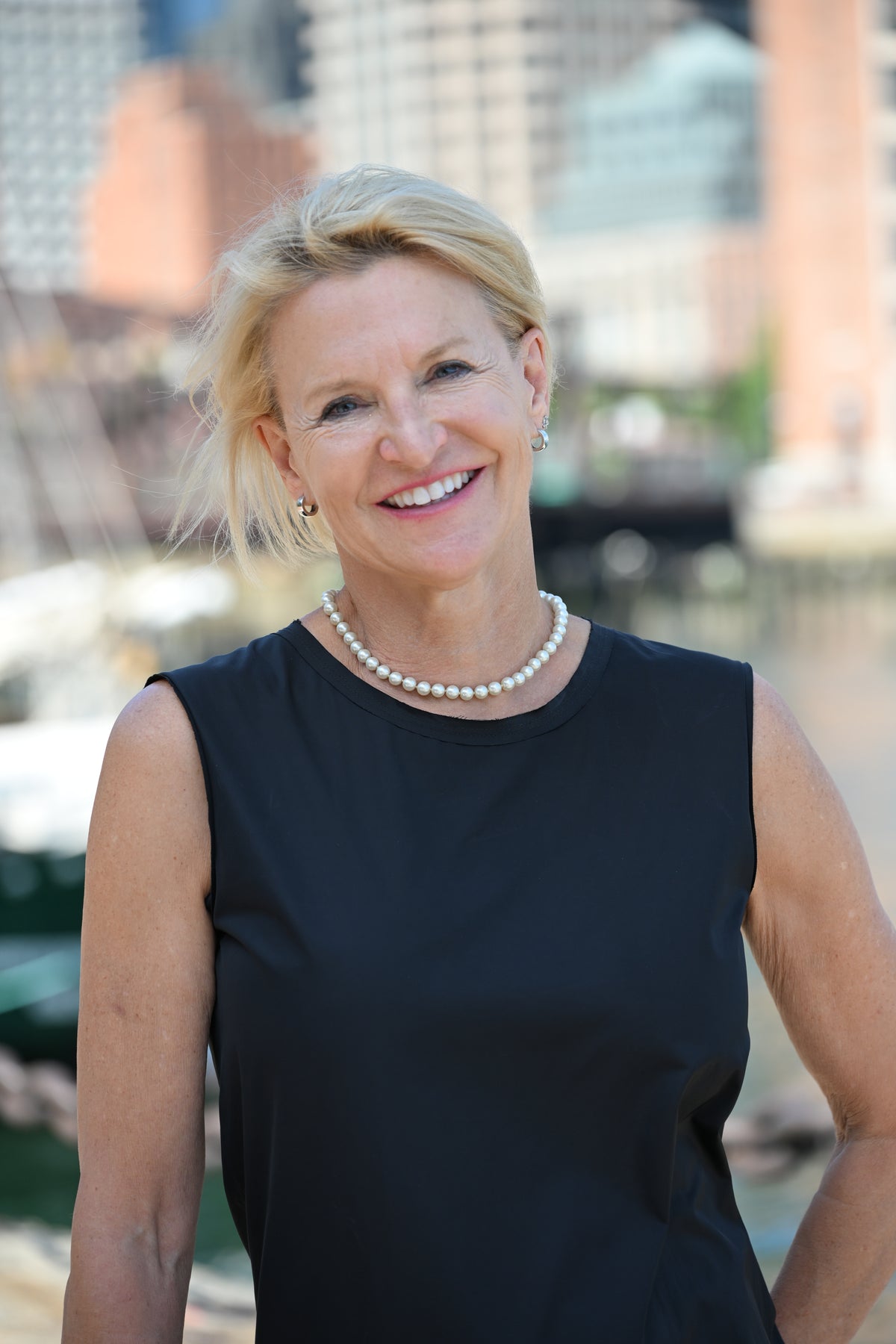 A smiling blonde woman wearing a sleeveless black dress and a pearl necklace, standing outdoors with a blurred city and water background.