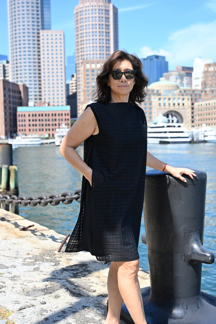 A woman wearing a black sleeveless PKTS Dana dress with a black Karami-ori cloth overlay—one hand through the side slit of the overlay and the deep, functional pocket—stands on a waterfront promenade with a city skyline and boats in the background.