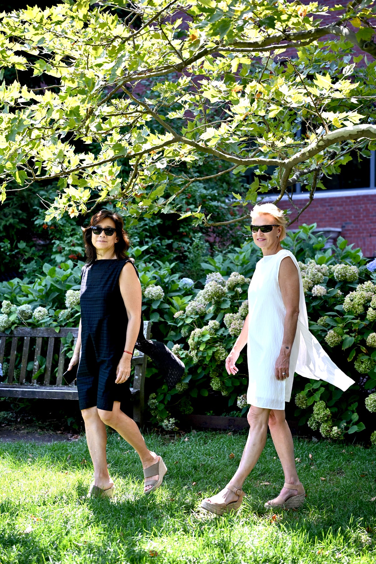 Two women walk outdoors on a grassy lawn under a leafy tree. The woman on the left wears a sleeveless black PKTS Dana dress with a Karami-ori cloth overlay. The woman on the right wears a white sleeveless PKTS Dana dress with a White Cloud overlay.
