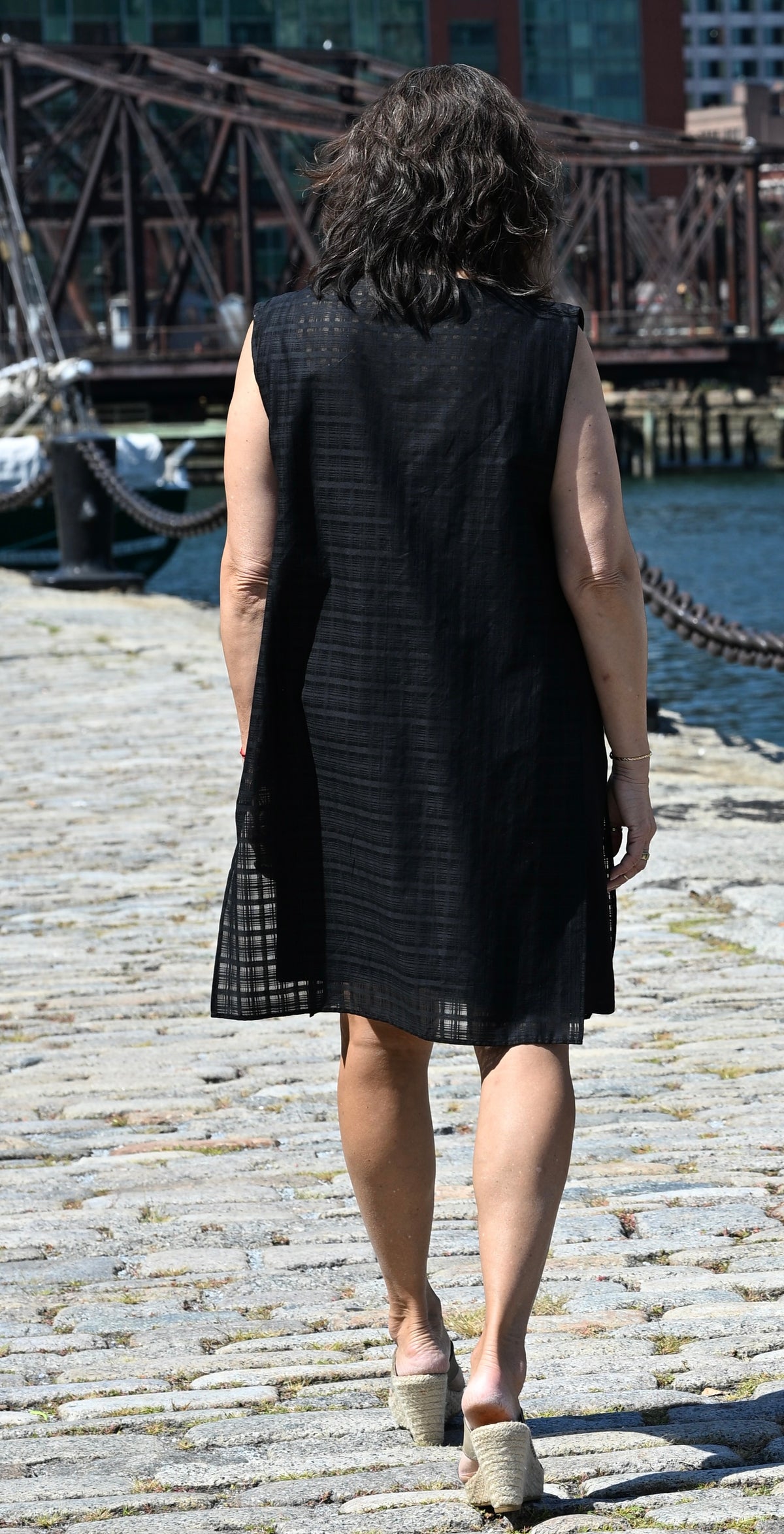 A woman viewed from behind, wearing a black sleeveless PKTS Dana dress with a Karami-ori cloth overlay, walks on a cobblestone dock next to water and a bridge.