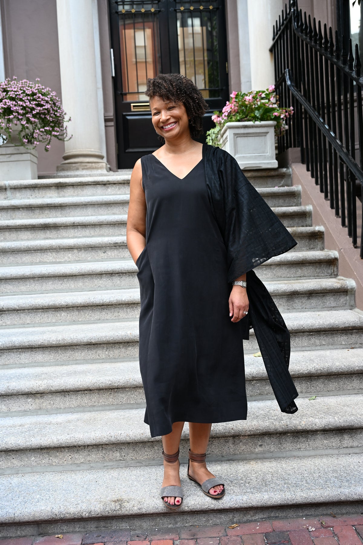 A woman stands on outdoor stone steps in front of a building entrance, wearing a black sleeveless PKTS Kelley midi dress with a Karami-ori cloth wrap on one sleeve.