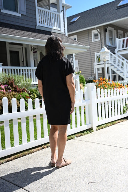 A woman viewed from behind wearing a black short-sleeve dress, standing on a concrete sidewalk in front of a white picket fence and residential houses.