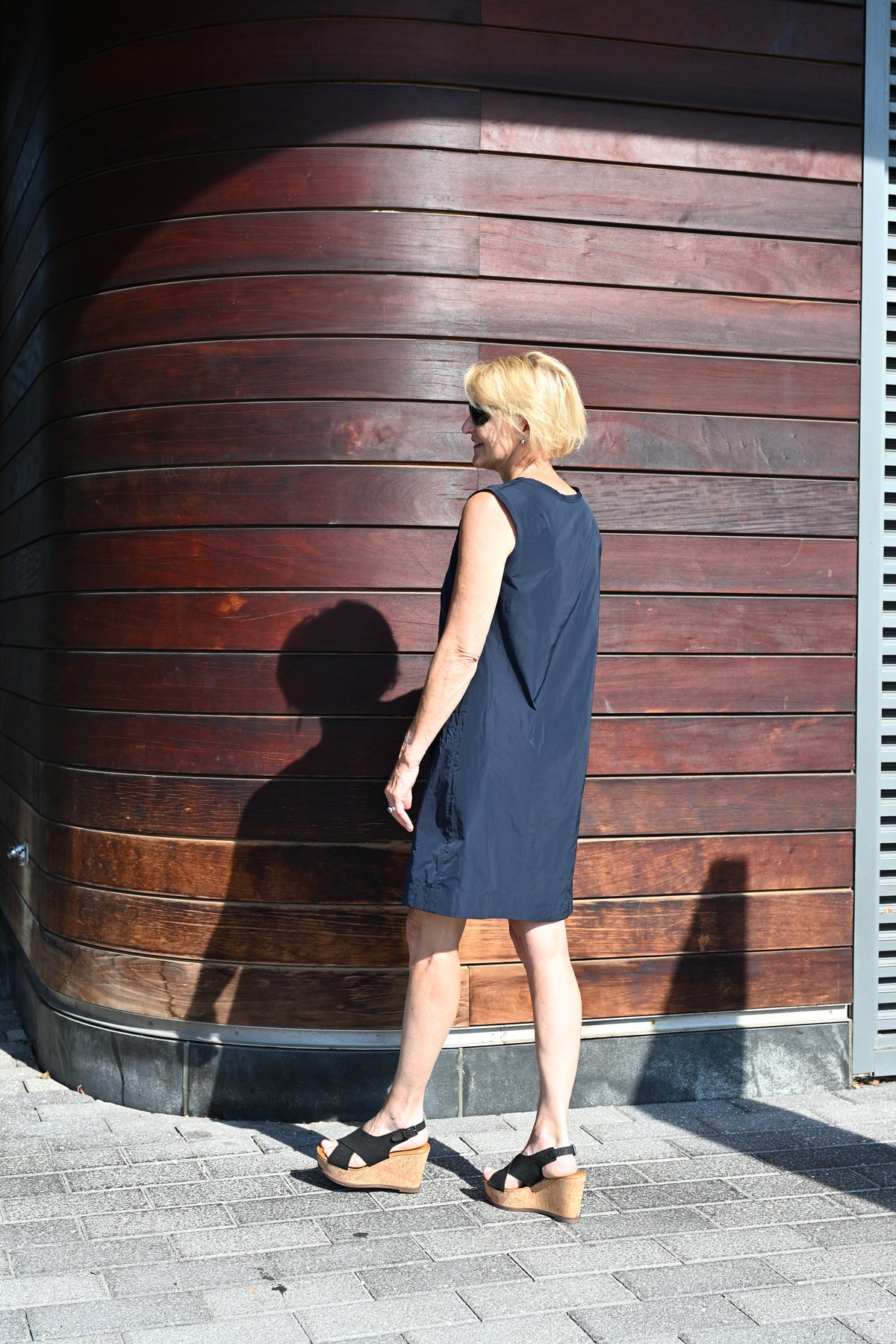 A woman with short blonde hair and sunglasses stands in profile wearing a sleeveless navy blue dress and black wedge sandals, posing in front of a curved dark brown wooden wall.