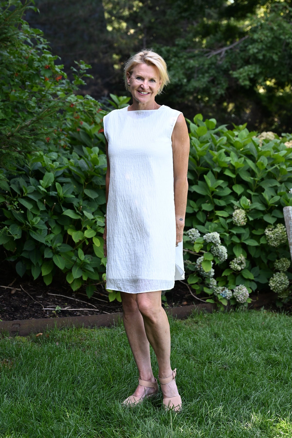 A woman wearing a sleeveless white PKTS Dana dress with a PKTS White Cloud overlay in a garden setting with lush green bushes and trees behind her.